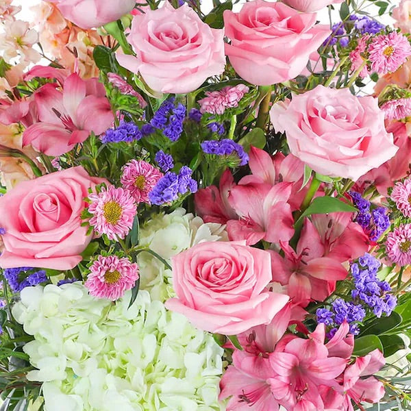 A floral arrangement featuring a variety of pink, purple, and white flowers with green foliage, arranged in a clear glass vase.