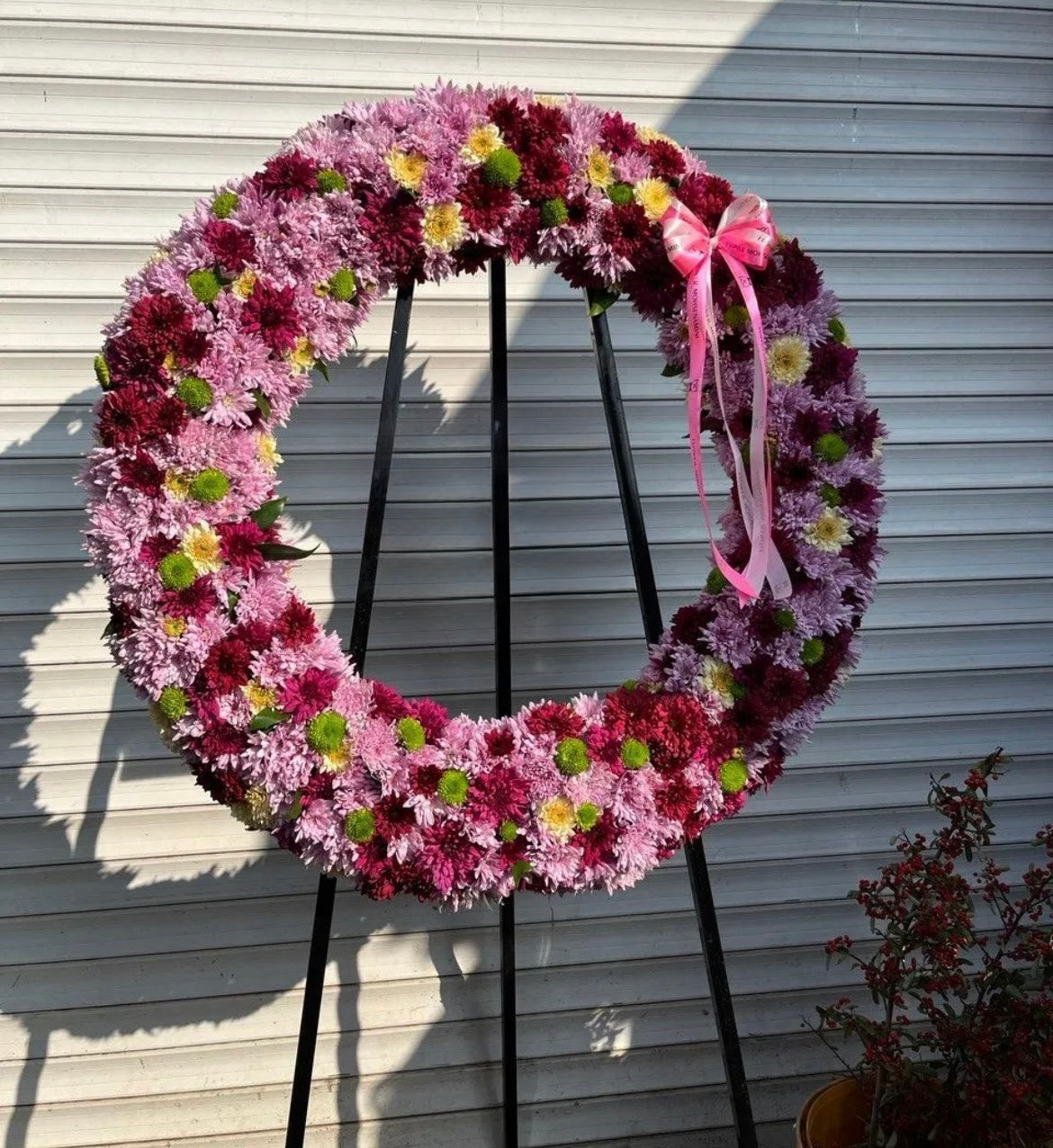 A large circular floral wreath made of pink, purple, and burgundy chrysanthemums with small green centers and a pink ribbon bow, displayed on a black metal stand against a white siding wall.