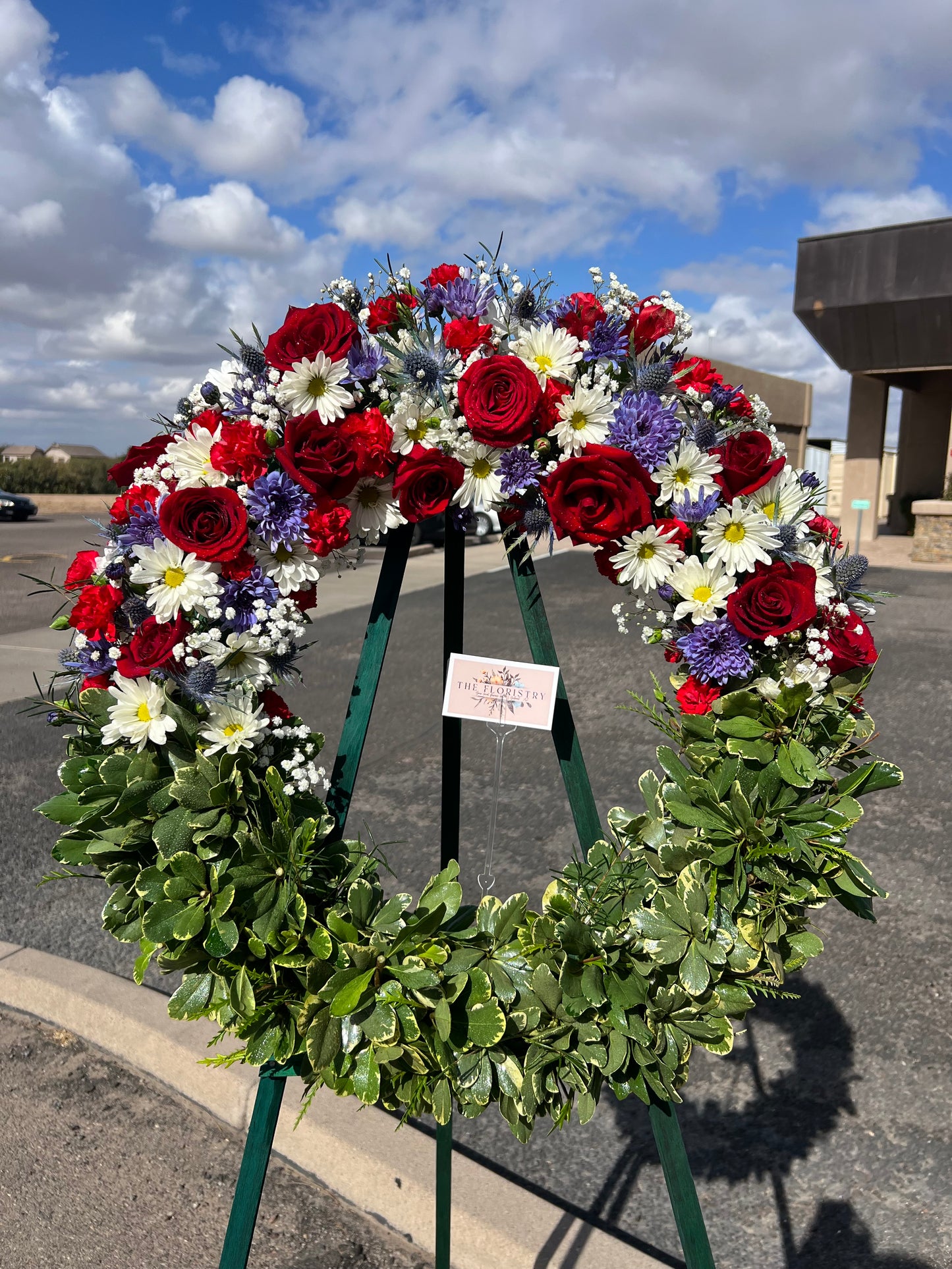 A large floral wreath with red roses, white daisies, purple flowers, and green foliage.