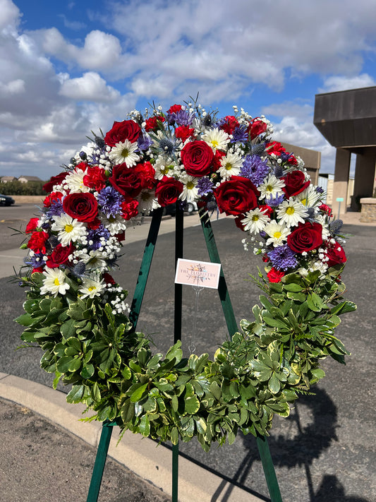 A large floral wreath with red roses, white daisies, purple flowers, and green foliage.