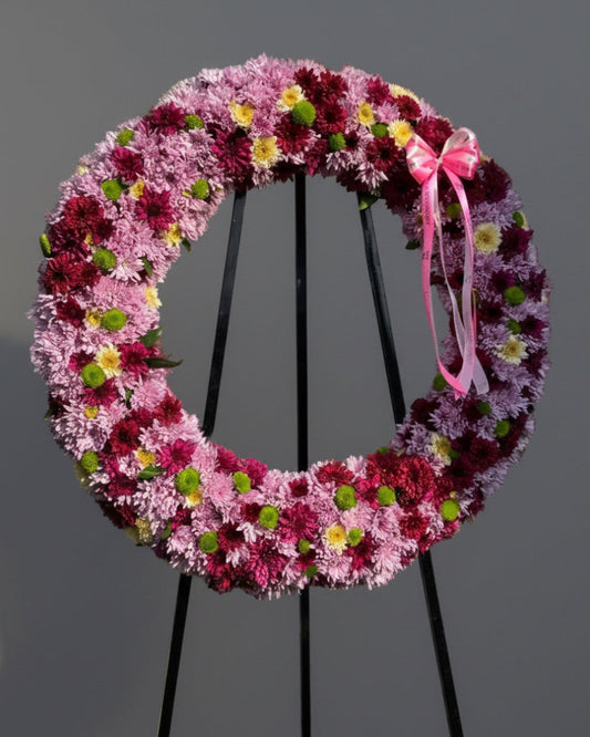 A large circular floral wreath made of pink, purple, and burgundy chrysanthemums with small green centers and a pink ribbon bow, displayed on a black metal stand against a white siding wall.